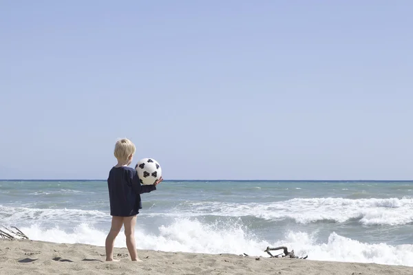 Malý chlapec s futball kouli stojící na pláži — Stock fotografie