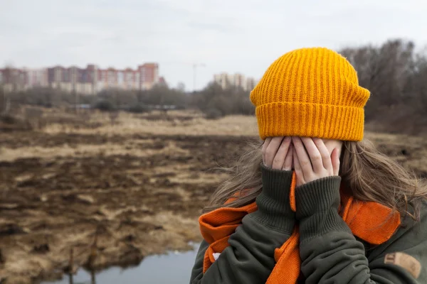Smutny, nastolatek pomarańczowy knitten kapelusz i szalik zamknięte twarz rękoma, samodzielny w pobliżu pole spalonej. Wiosna — Zdjęcie stockowe