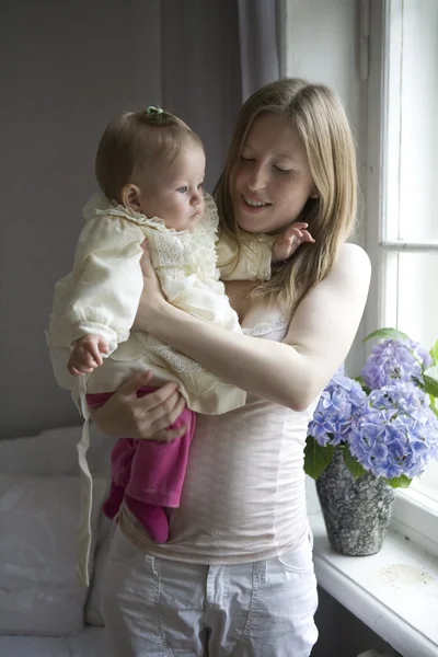 Moeder houdt haar blond glimlachende babymeisje permanent ter venster — Stockfoto