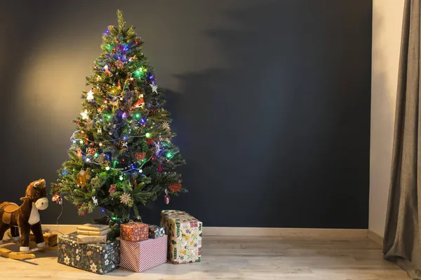 房间里矗立着一棵装饰着花环和木制玩具的圣诞树 背景是深蓝色的 树下有几箱礼物和一匹摇曳的马 复制空间 — 图库照片