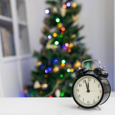 Beyaz bir masadaki siyah bir çalar saat saat on ikiyi gösteriyor. Arka planda odakta olmayan bir Noel ağacı var..