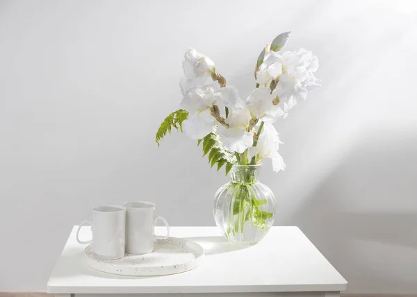 テーブルの上の透明な花瓶に3つの白い虹彩とシダの花束 トレイに2つのセラミックティーカップ — ストック写真
