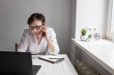 Orta yaşlı, kısa saçlı, gözlüklü esmer bir kadın pencerenin yanındaki dizüstü bilgisayarda çalışıyor. Ofis çalışanı telefonda konuşuyor ve gülüyor.