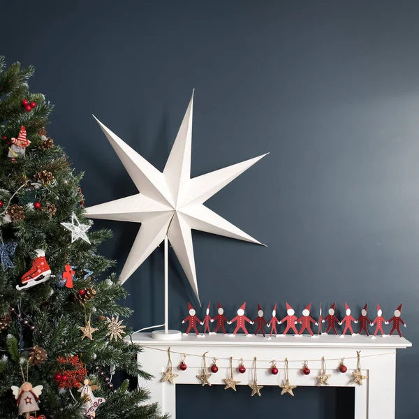 白い紙を手にした赤い紙の男性と木の背景にある大きな紙の星クリスマスの内装として濃い青の壁の背景に人工暖炉があります — ストック写真