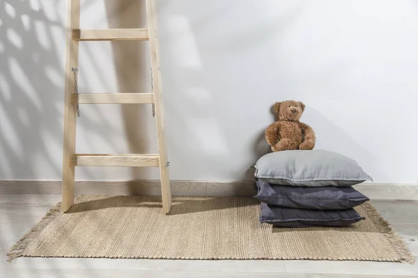 The teddy bear is leaning against the wall next to a small children\'s stool with three gray pillows on it. The stepladder is leaning against the wall