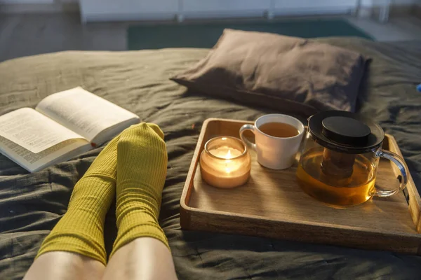 ベッドの上の毛むくじゃらの靴下の女性の足の柔らかい写真とトレイの上にお茶とキャンドルの本とカップ インテリアと家庭の化粧品のコンセプト トップ表示 — ストック写真