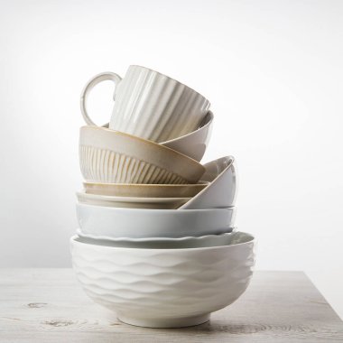 Mutfaktaki bej bir masada farklı boylarda ve renklerde beyaz kaseler, tabaklar ve bardaklar piramidi. İskandinav tarzı. Mesajın için yer aç. Boşluğu kopyala