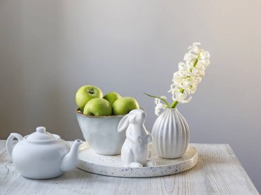 70 'li yılların tarzında flüt vazosundaki beyaz bir sümbül büyük bir kase yeşil elma, Paskalya porseleni ve bir çaydanlık masanın üzerinde. Minimalizm. İskandinav tarzı. Metin için yer
