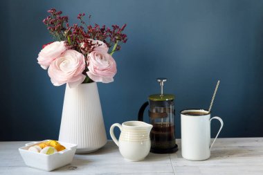 Kahve fincanı ve bir buket pembe İran düğün çiçeği, ahşap masada. Tasarım için arkaplan oluşturun. Beyaz bir porselen kasede sarı ve beyaz badem kurabiyeleri. Koyu mavi arka plan. Boşluğu kopyala