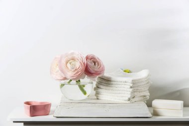 Şeffaf yuvarlak vazoda üç soluk pembe ranunculus ve beyaz pencere pervazında mumlar. Yuvarlak el ve yüz havlusu. Boşluğu kopyala. Metin için yer
