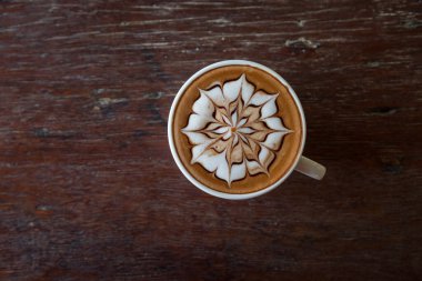 Üzerinde latte resmi olan bir fincan kahve ve ahşap masa arkası.