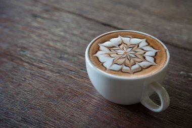 Üzerinde latte resmi olan bir fincan kahve ve ahşap masa arkası.