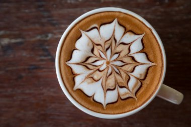 Üzerinde latte resmi olan bir fincan kahve ve ahşap masa arkası.
