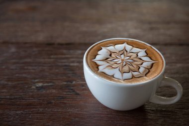 Üzerinde latte resmi olan bir fincan kahve ve ahşap masa arkası.