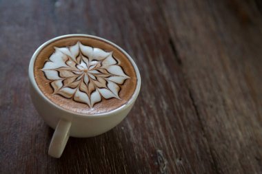 Üzerinde latte resmi olan bir fincan kahve ve ahşap masa arkası.