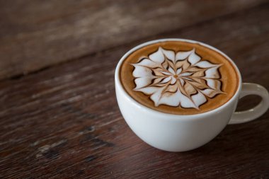 Üzerinde latte resmi olan bir fincan kahve ve ahşap masa arkası.