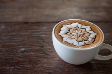 Üzerinde latte resmi olan bir fincan kahve ve ahşap masa arkası.