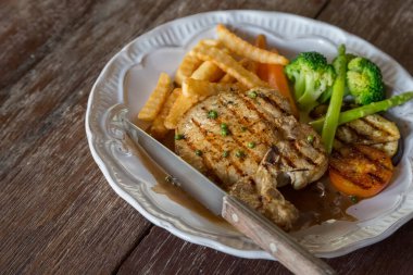 Ahşap masa arkasında beyaz tabakta siyah biberli domuz pirzolası.