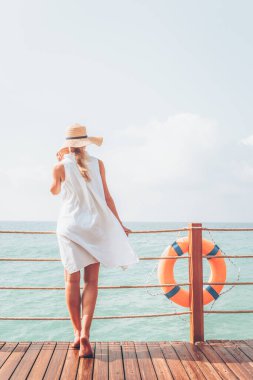 Beyaz elbiseli bronz tenli kadın ahşap iskelede deniz manzarasını sever.