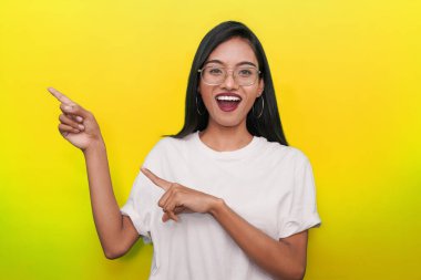 Sarı arka planda beyaz tişört giyen genç Asyalı kadın sol tarafını işaret ediyor..