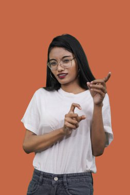 Beyaz tişörtlü, turuncu arka planda genç Asyalı kadın utangaçlıkla işaret ediyor.