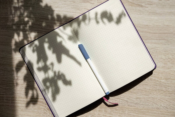 Mock-up of notebook with clean beige sheets and pen for writing, on wooden table in rays of sunlight. Sun's rays, through branches of trees, fall on surface of object, casting hard shadow.
