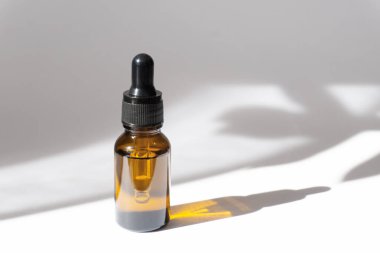 Beyaz arka planda kahverengi camdan yapılmış damlalık şişesi. Ağaç dalının sert gölgesi masanın yüzeyine düşer. Doğal kozmetik, vücut bakımı, aromaterapi kavramı.