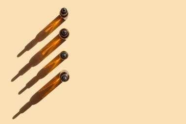 Dört adet damlalık kapaklı cam şişe, izole edilmiş bej arka planda sıraya dizilmiş. Yüzeyde sert gölgeler bırakan nesnelerin üst görünümü. Güzellik endüstrisi kavramı.