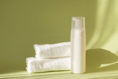 Üzerinde şeker kutusu olan beyaz bir şişe ve yeşil arka planda iki terry havlusu, güneş ışığı ışınlarıyla. Kozmetik losyon için ambalaj, yüz kaplaması için köpük temizleme.