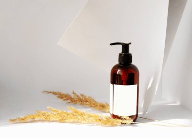 Kozmetik için bir şişe viski ve kuru ot dalları arasında boş bir etiket. Nesnelerin üzerine düşen güneş ışığıyla beyaz bir zemin. Güzellik endüstrisi.