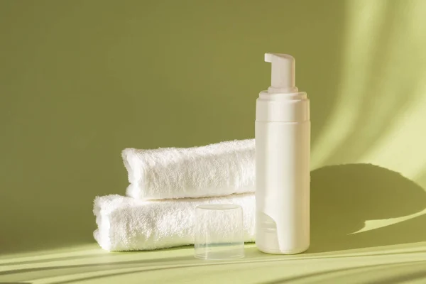 Mock-up of an open white bottle with dispenser and two terry towels on green background, in rays of sunlight. Concept of natural cosmetics, care for body, face.