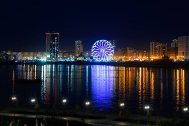 Nehir kıyısındaki yanan binalar ve fenerler, gece şehrinin çok renkli ışıkları suya yansıdı. Toprak zeminde dönme dolap..
