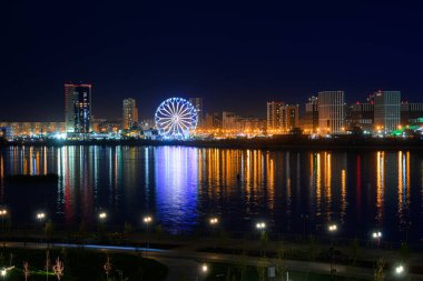 Nehir kıyısındaki yanan binalar ve fenerler, gece şehrinin çok renkli ışıkları suya yansıdı. Toprak zeminde dönme dolap..