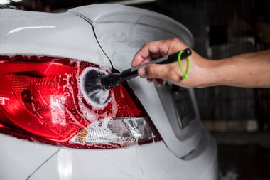 Bir adam araba şampuanıyla kaplı beyaz bir sedanın arka lambasını güzel bir fırçayla temizliyor. Araba yıkamada veya oto yıkamada hizmet verilen bir araç.