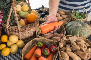 Bir erkek satın almak için büyük ve dolgun bir havuç seçer. Arka planda çeşitli sebzeler ve meyveler var. Küçük bir marketten alışveriş..