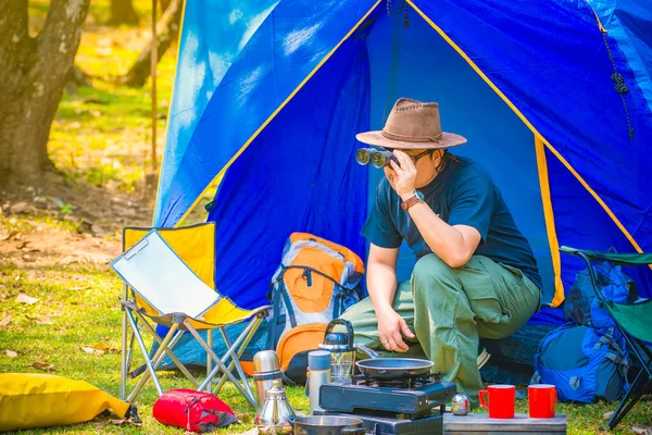 A man at the camping area. He is using binoculars with his notepad on a yellow chair. there are have an outdoor kitchen equipment and lantern, mug and tent.