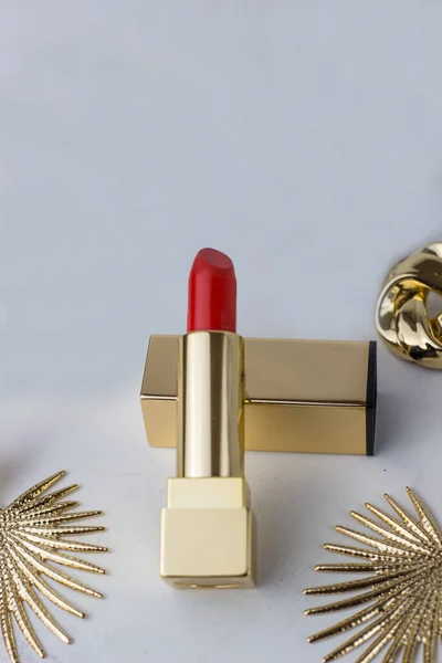 Red lipstick in a gold case on a white background surrounded by gold earrings