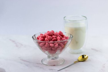 Beyaz mermer arka planda bir kase kahvaltı gevreği ve bir bardak süt. Ahududu aromalı atıştırmalıklar.