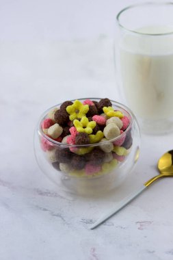 Top şeklinde bir kase abur cubur, beyaz mermer bir masada kalpler ve çiçekler. Kahvaltı gevreği ve bir bardak süt.