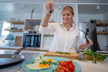 Modern bir mutfakta yemek pişiren güzel genç bir kadın. Tavada kızarmış yumurtanın üzerine doğranmış taze soğan serpin. Evde yalnızım. Seçici odak.