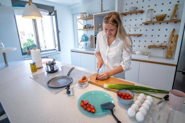 Modern bir mutfakta yemek pişiren güzel genç bir kadın. Kirazlı domatesi ikiye bölmek, yumurtalı kahvaltı hazırlamak. Evde yalnızım, tek kişilik yemek yapıyorum..