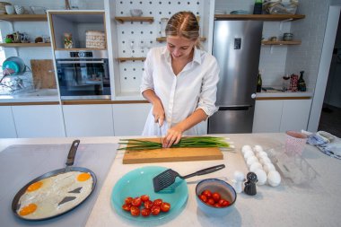 Modern bir mutfakta yemek pişiren güzel genç bir kadın. Taze soğan dilimlemek, yumurtalı kahvaltı hazırlamak. Evde yalnızım, tek kişilik yemek yapıyorum..