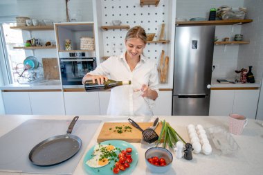 Modern donanımlı bir mutfakta yemek pişiren güzel sarışın kadın. Kızarmış yumurta hazırlamak ve bir bardağa şarap dökmek. Kahvaltı mı yoksa akşam yemeği mi?.