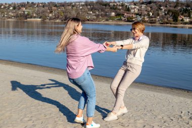 Yetişkin kızıyla kaynaşan ve nehir kenarında eğlenen mutlu kıdemli anne. El ele tutuşup kumsalda dans ediyorlar..