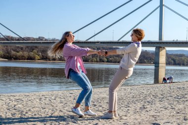Yetişkin kızıyla kaynaşan ve nehir kenarında eğlenen mutlu kıdemli anne. El ele tutuşup kumsalda dans ediyorlar..