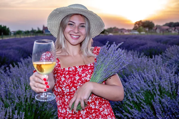 Portrait of a young beautiful woman toasting a glass of wine in a lavender field at scenic summer sunset.