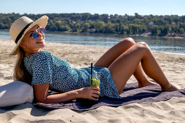 Beautiful young woman with a straw hat, lying on a deserted sandy beach, enjoying the sun and cocktail.