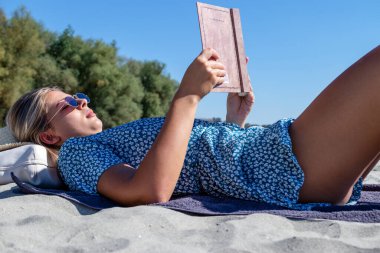 A beautiful young woman with sunglasses reading a book, and enjoying her time alone on a deserted sandy beach. 