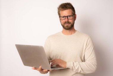 Süveter giyen, beyaz arka planda dizüstü bilgisayarı olan gülümseyen genç bir adamın stüdyo portresi.