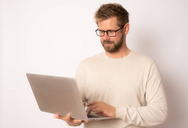 Süveter giyen, beyaz arka planda dizüstü bilgisayarı olan gülümseyen genç bir adamın stüdyo portresi.